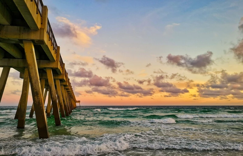 a view of the ocean under a pier at sunset, by Robbie Trevino, renaissance, sunset with cloudy skies, the emerald coast, in a golden sunset sky, towering waves