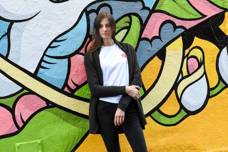 a woman standing in front of a colorful wall, inspired by Christen Dalsgaard, graffiti, outlive streetwear collection, cardigan, portrait of alexandra daddario, dressed in a white t shirt