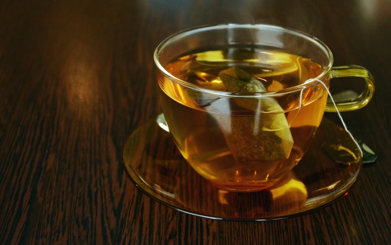 a cup of tea sitting on top of a wooden table, by Aleksander Gierymski, pixabay, green tea, glass refraction, “ golden cup, olive oil