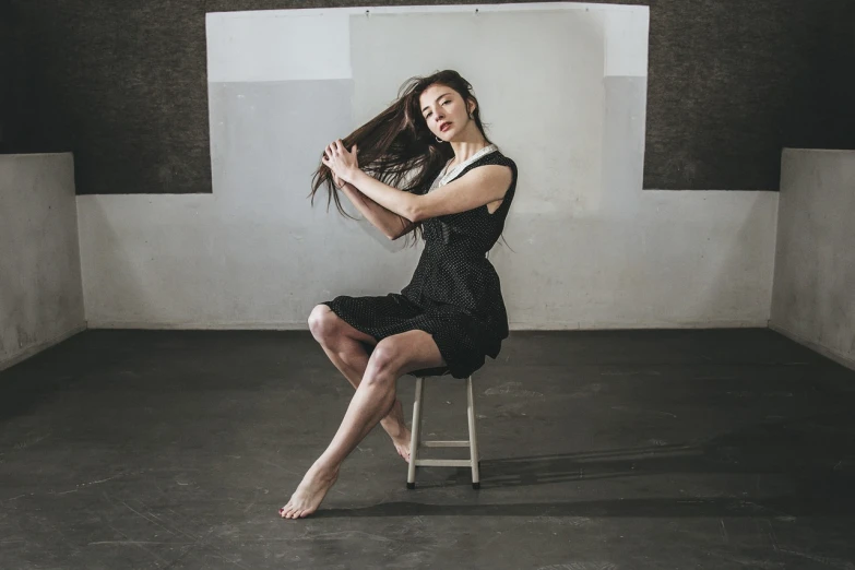 a woman sitting on a chair in a room, a portrait, by Elizabeth Polunin, art photography, contemporary dance poses, young woman with long dark hair, she is wearing a black dress, sitting on a stool