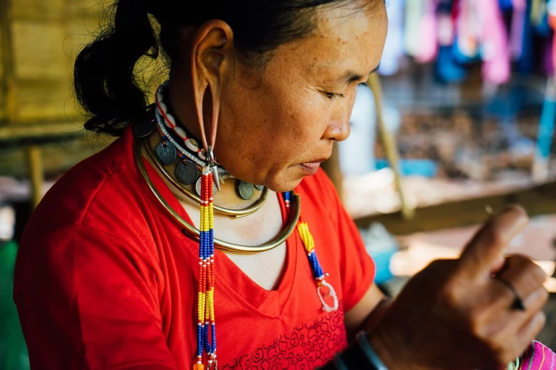 a woman in a red shirt looking at a cell phone, by Edwin Georgi, pexels contest winner, cloisonnism, tribal jewelry, long neck, one of the weavers of destiny, beads