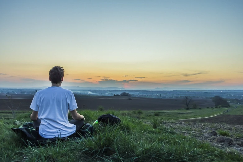 a man sitting on top of a lush green hillside, by Julian Allen, pexels, hindu stages of meditation, evening at dusk, looking onto the horizon, featured