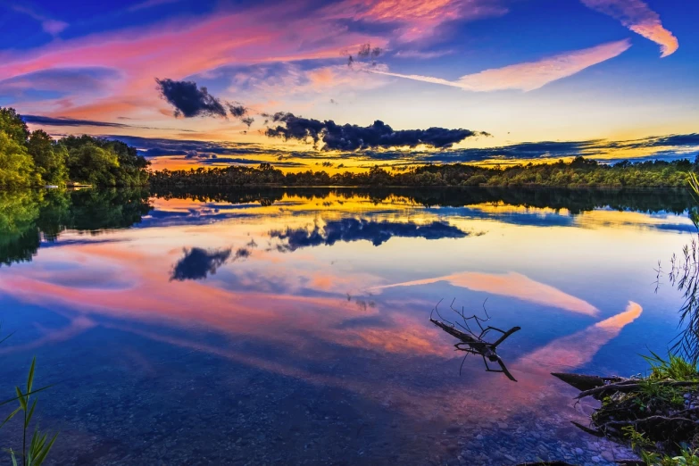 a large body of water next to a forest, a picture, by Antoni Brodowski, shutterstock, violet and yellow sunset, blue and pink colors, mirror reflection, colorful clouds