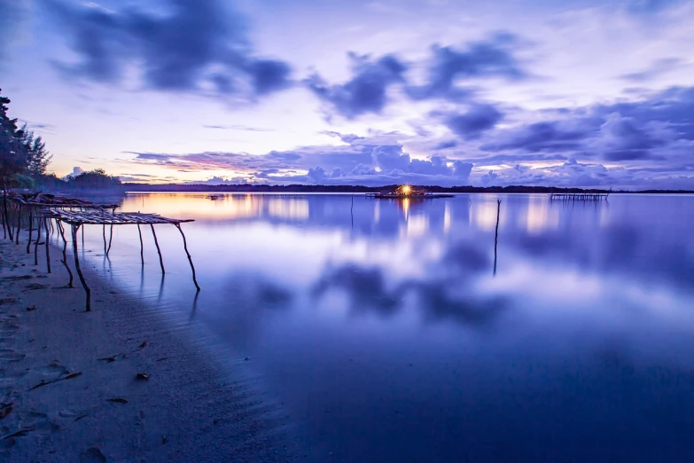 a group of chairs sitting on top of a beach next to a body of water, a picture, by Niklaus Manuel, blue hour photography, water reflection!!!!!, at purple sunset, calm clouds