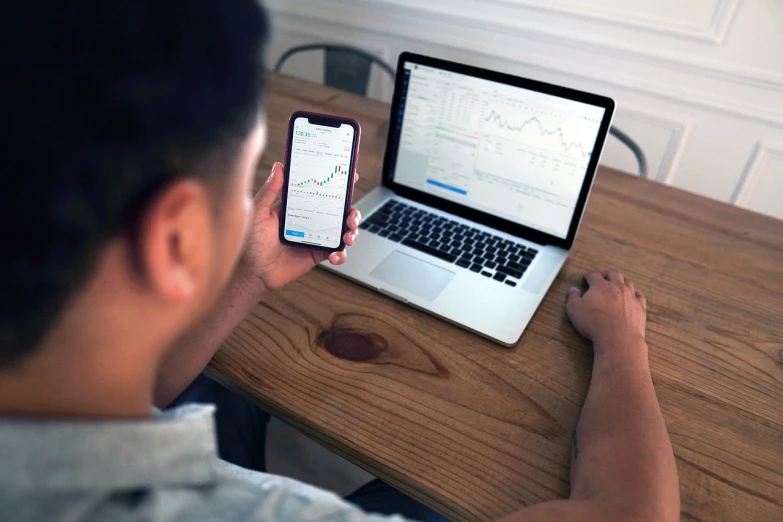 a man sitting at a table with a laptop and a cell phone, a picture, by Adam Rex, pexels, dau-al-set, displaying stock charts, square, crypto, clean background trending