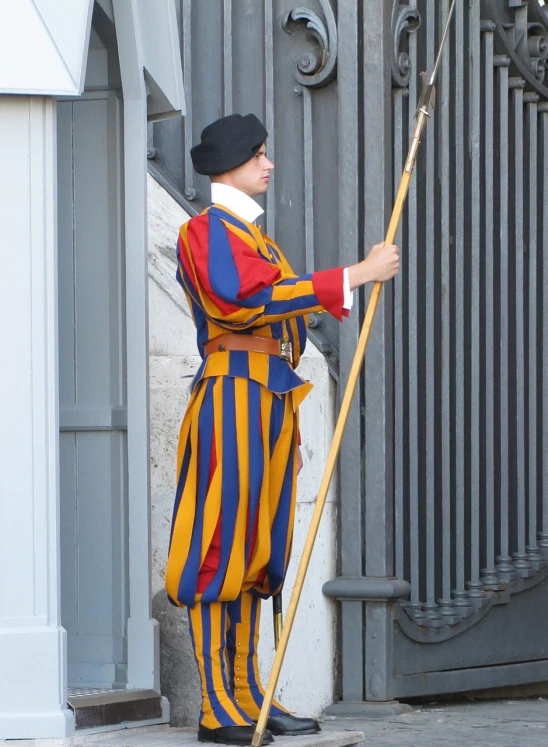 a man that is standing in front of a gate, inspired by Cagnaccio di San Pietro, colorful uniforms, stripes, wooden staff, parliament