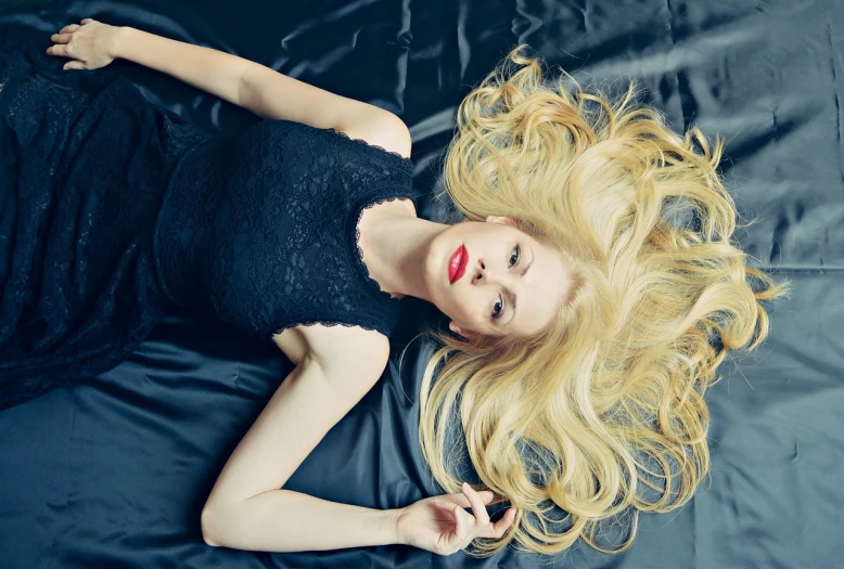 a woman with blonde hair laying on a bed, by Zofia Stryjenska, tumblr, she is wearing a black dress, floating long hair, red lips, top down photo