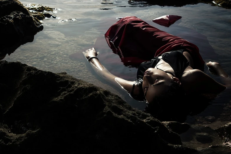 a woman laying on top of a rock next to a body of water, a photo, inspired by Brooke Shaden, romanticism, trent parke, crime scene photography, hot sun, blood in the seahighly