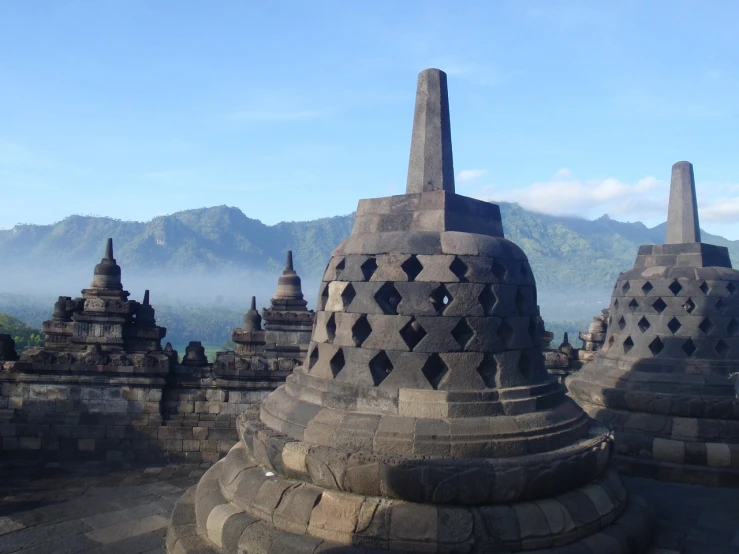 a number of stone structures with mountains in the background, a picture, flickr, sumatraism, black domes and spires, statue of a cubes and rings, morning lighting, cone