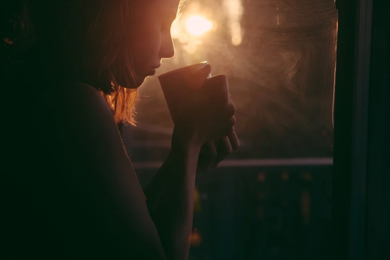 a woman holding a cup in front of a window, a picture, inspired by Elsa Bleda, pexels, romanticism, in a sunset haze, on a dark background, backlit face, coffee smell