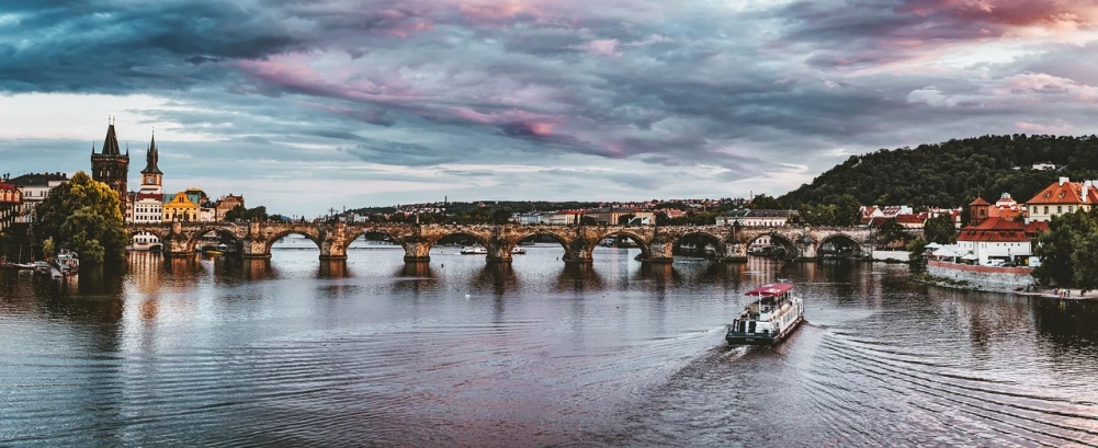 a boat traveling down a river next to a bridge, by Matija Jama, pexels contest winner, art nouveau, pink and grey clouds, prague, panoramic, 1647