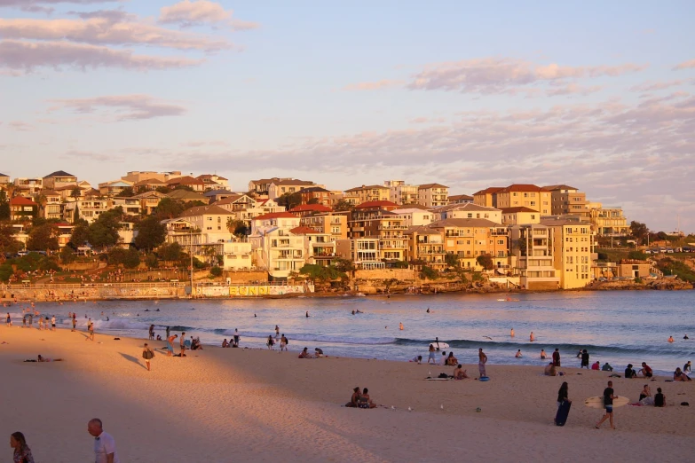 a group of people standing on top of a sandy beach, a picture, by Thomas Baines, shutterstock, bondi beach in the background, waterfront houses, soft golden light, telephoto vacation picture