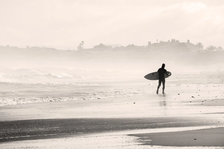 a man standing on top of a beach holding a surfboard, a black and white photo, shutterstock, misty weather, walking to the right, soft colors mono chromatic, action sports photography