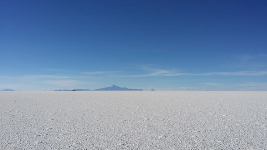 a snow covered field with a mountain in the distance, a picture, by Andrei Kolkoutine, unsplash, minimalism, constructed upon salar de uyuni, giant crater in distance, clear blue sky, white salt