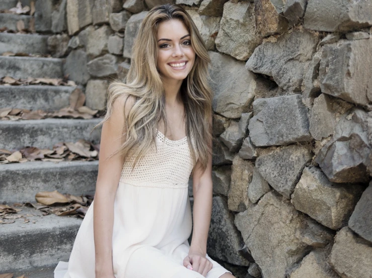 a beautiful young woman sitting in front of a stone wall, by Amelia Peláez, happening, cute smile, flowing blonde hair, dreamy mila kunis, dslr photo of a pretty teen girl