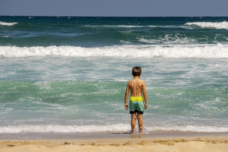 a young boy standing on top of a beach next to the ocean, by Etienne Delessert, pexels, figuration libre, green swimsuit, waves and splashes, on a hot australian day, stock photo