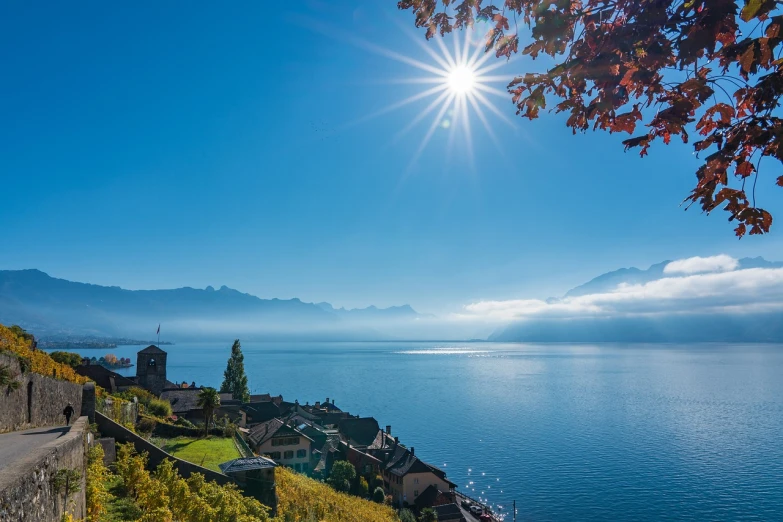 a large body of water sitting next to a lush green hillside, a stock photo, by Johannes Martini, shutterstock, sunbeams blue sky, switzerland, autumn season, village in the background