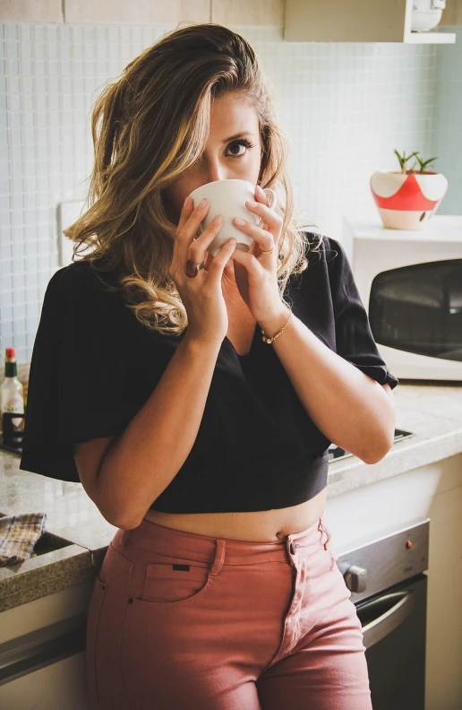 a woman standing in a kitchen holding a cup, by Sydney Carline, pexels, wearing a sexy cropped top, pink and black, is ((drinking a cup of tea)), blonde beautiful young woman