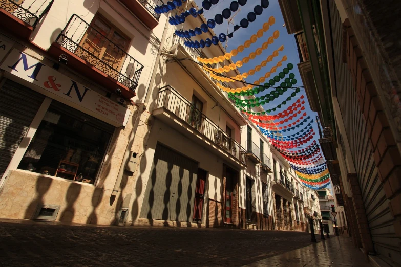 a group of people walking down a street next to tall buildings, by Pedro Álvarez Castelló, flickr, folk art, costa blanca, streamers, old town, the sun shines in