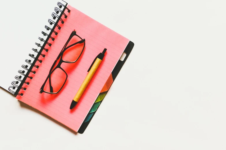 a notebook with a pen and glasses on top of it, a photo, minimalism, pink and red colors, high quality product photo