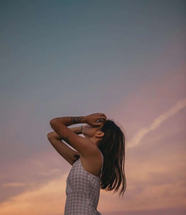 a woman standing on top of a beach next to the ocean, a picture, inspired by Elsa Bleda, aestheticism, sunset halo behind her head, hands in her hair. side-view, depressed girl portrait, pink sky