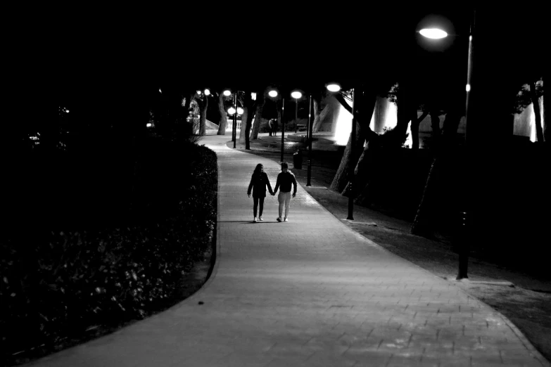 two people walking down a sidewalk at night, a black and white photo, by Alexis Grimou, pexels, in a park, lovers, alone!!, boardwalk