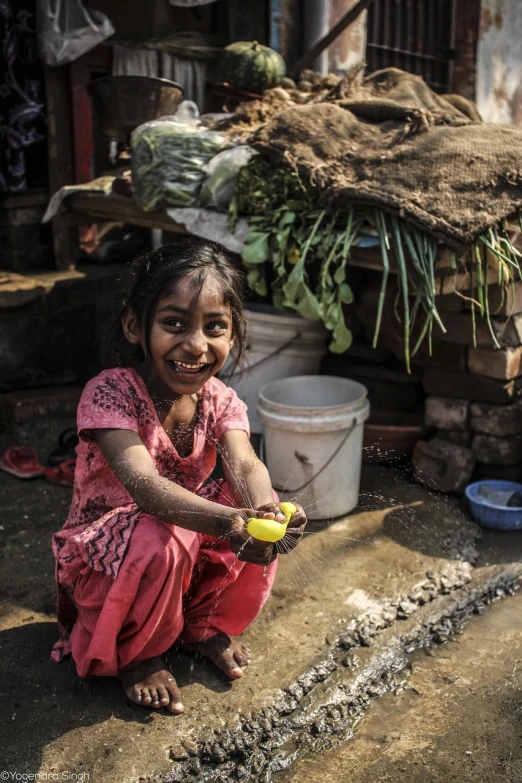 a little girl sitting on the ground holding a banana, a picture, by Jakob Gauermann, on an indian street, playing with the water, clean bright happy, uhd candid photo of dirty