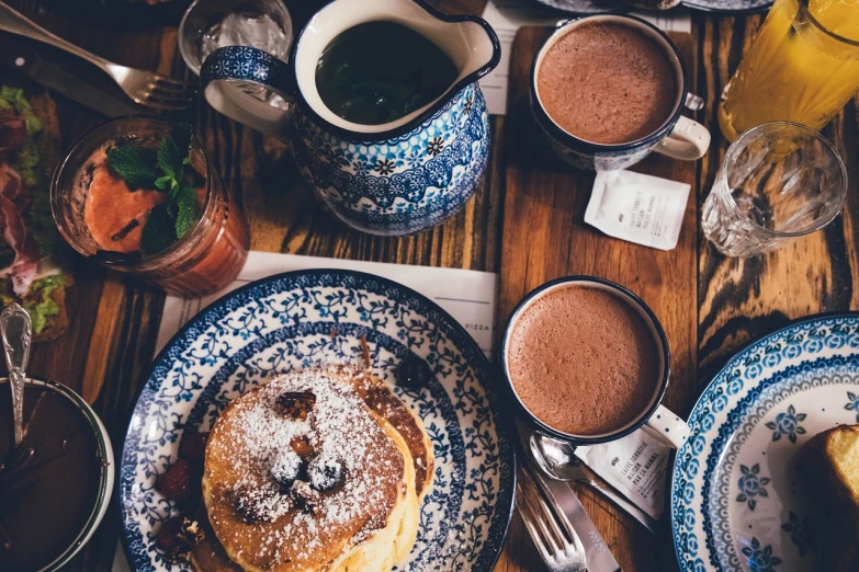 a table topped with plates of food and cups of coffee, a still life, by Lee Loughridge, pexels, living food adorable pancake, hot cocoa drink, blue mood, 🐝👗👾