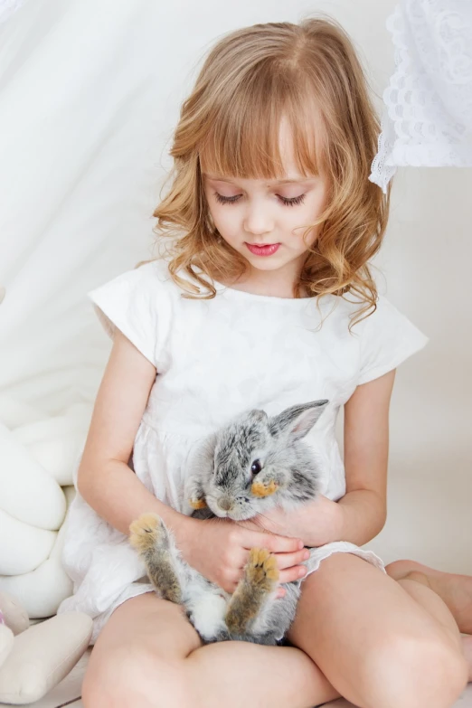 a little girl sitting on a bed holding a rabbit, a picture, by Nina Petrovna Valetova, shutterstock, romanticism, glossy white, stock photo, blond furr, closeup photo