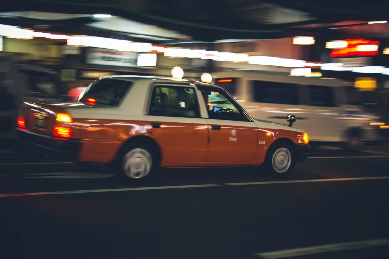 a taxi driving down a city street at night, a tilt shift photo, unsplash, orange and white color scheme, vintage footage on tokyo streets, left profile, low angle photo