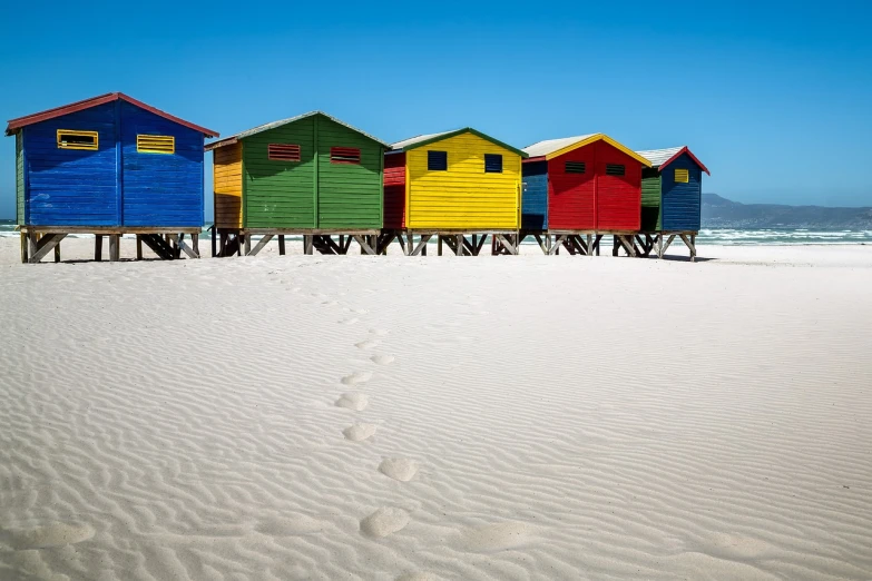 a row of colorful beach huts sitting on top of a sandy beach, a picture, by Randy Post, shutterstock, fine art, south african coast, full of colour 8-w 1024, vibrant deep saturated colors, an ancient