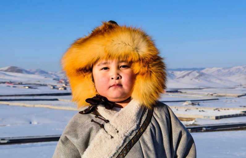 a person standing in the snow wearing a furry hat, a portrait, hurufiyya, am a naranbaatar ganbold, boy with neutral face, sky-blue thick fur robes, amber