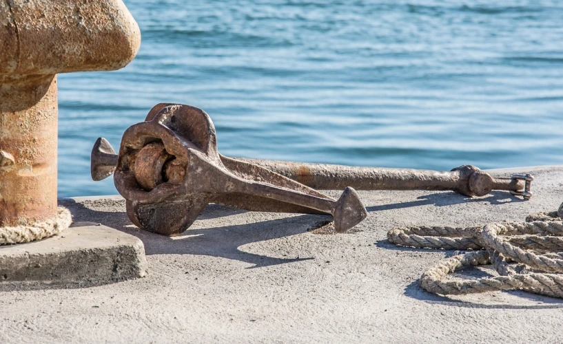 a large anchor sitting on top of a dock next to a body of water, a portrait, by Raymond Normand, shutterstock, figuration libre, many rusty joints, very sharp and detailed image, levers, resting