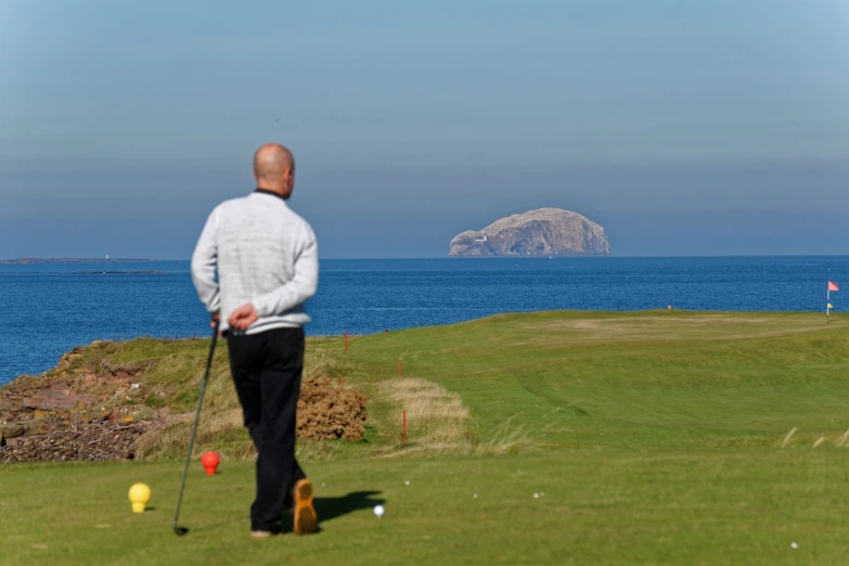 a man standing on top of a lush green field, by John Murdoch, flickr, golf course in background, looking out over the sea, biggish nose, blonde