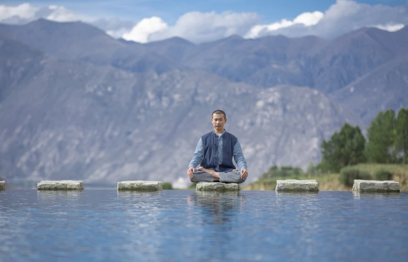 a man sitting in the middle of a body of water, a portrait, sitting cross-legged, with mountains in the background, shodan, yang qi
