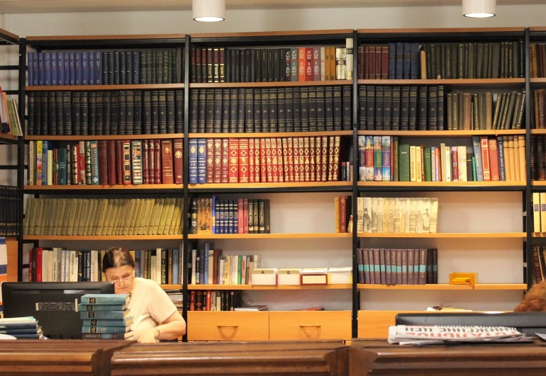 a woman sitting at a desk in a library, a photo, by Erwin Bowien, orthodox, panoramic shot, shelves full, blog-photo