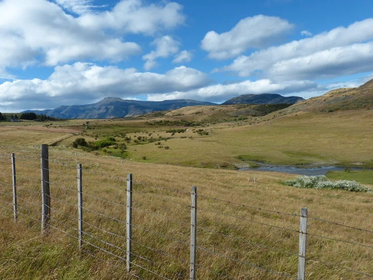 a field with a fence and mountains in the background, a picture, by Colin Middleton, flickr, hurufiyya, kahikatea, stream, blonde, highlands