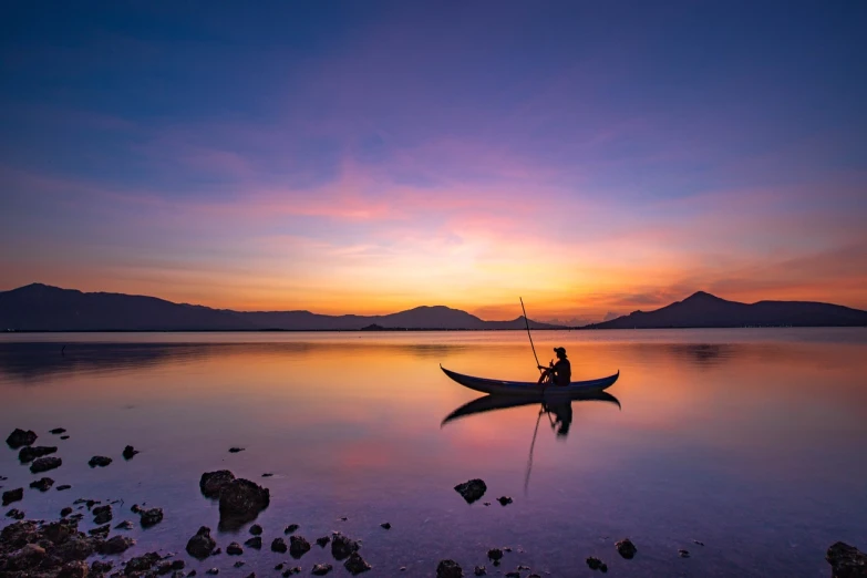 a man sitting in a boat on a lake at sunset, a picture, by Basuki Abdullah, vibrant vivid colors, istock, blue sunset, violet and yellow sunset