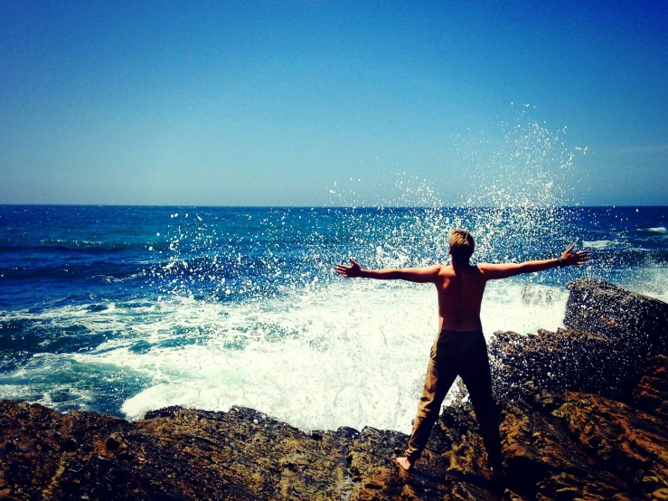 a man standing on top of a rock next to the ocean, a picture, splashing, pose(arms up + happy), ocean spray, katey truhn