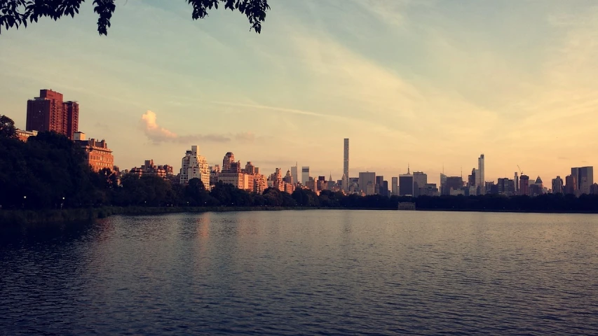 a large body of water with a city in the background, a picture, by Niko Henrichon, central park, sun rise, phone photo, tastes