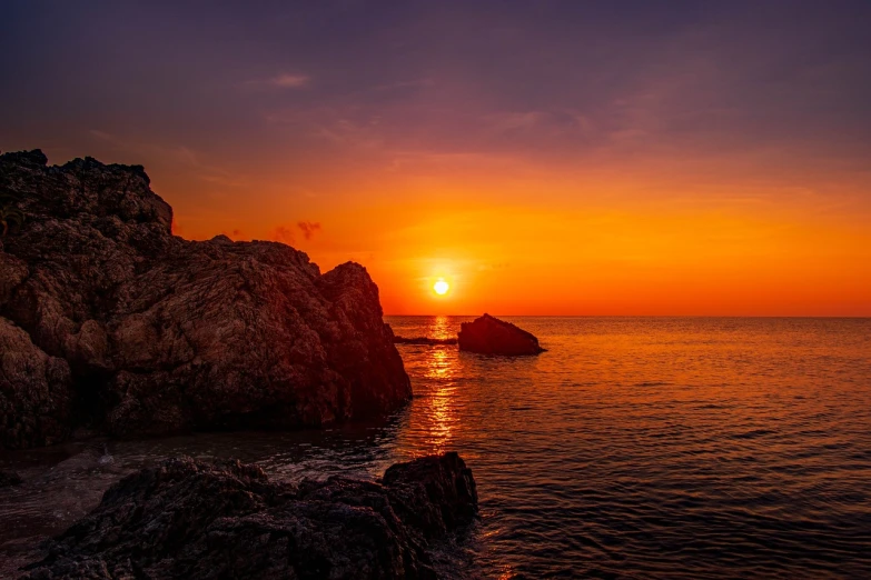 a sunset over the ocean with rocks in the foreground, a picture, by Alexis Grimou, pexels, romanticism, red sun over paradise, deep purple and orange, costa blanca, 8k hdr sunset lit