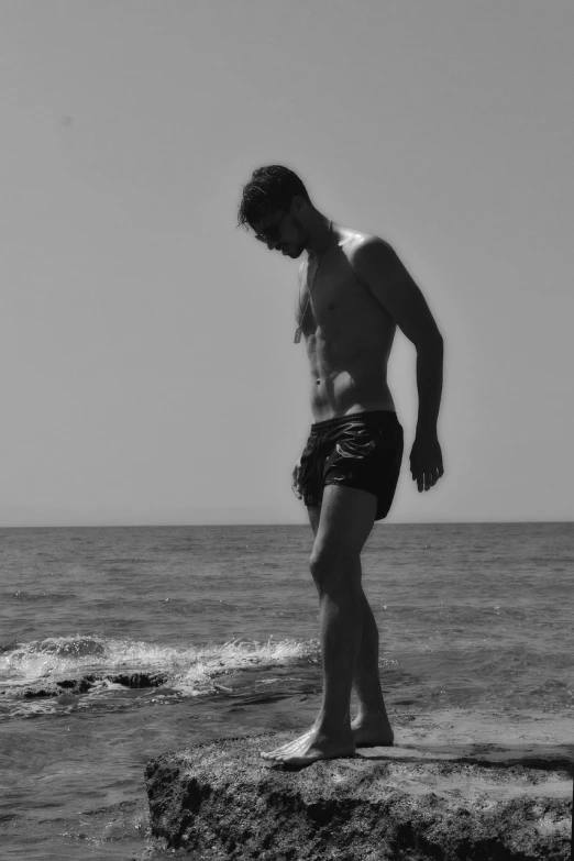 a man standing on top of a rock next to the ocean, a black and white photo, by Alexis Grimou, mid-shot of a hunky, christian dimitrov, 3/4 side view, character is half in the water