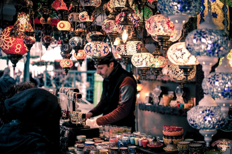 a man that is standing in front of a counter, a tilt shift photo, pexels, cloisonnism, colorful lanterns, middle eastern style vendors, moody lights!! intricate, holiday season