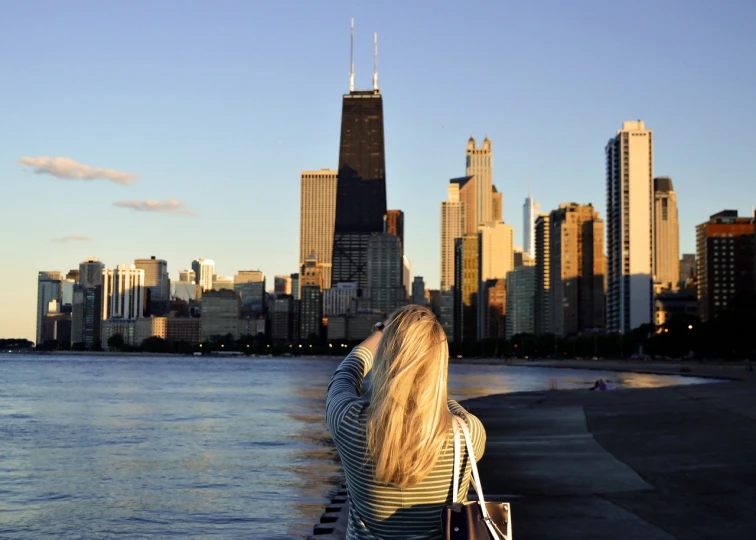 a woman standing next to a body of water with a city in the background, a photo, by Whitney Sherman, chicago skyline, blonde, girl watching sunset, usa-sep 20