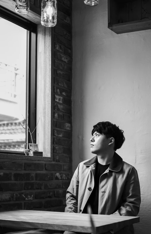 a man sitting at a table in front of a window, a picture, inspired by Kim Myeong-guk, unsplash, he is wearing a black trenchcoat, bw photo, declan mckenna, window light