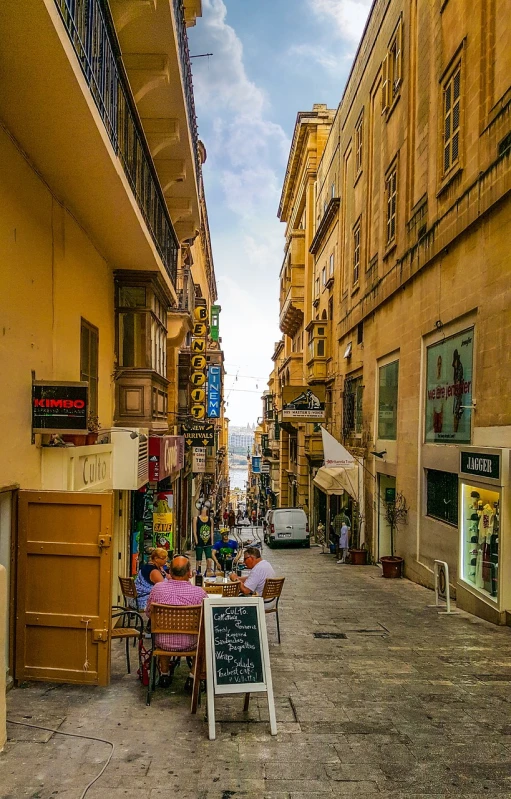 a couple of people sitting at a table in the middle of a street, a photo, by Simon Marmion, shutterstock, mediterranean vista, narrow footpath, dingy city street, outdoors tropical cityscape