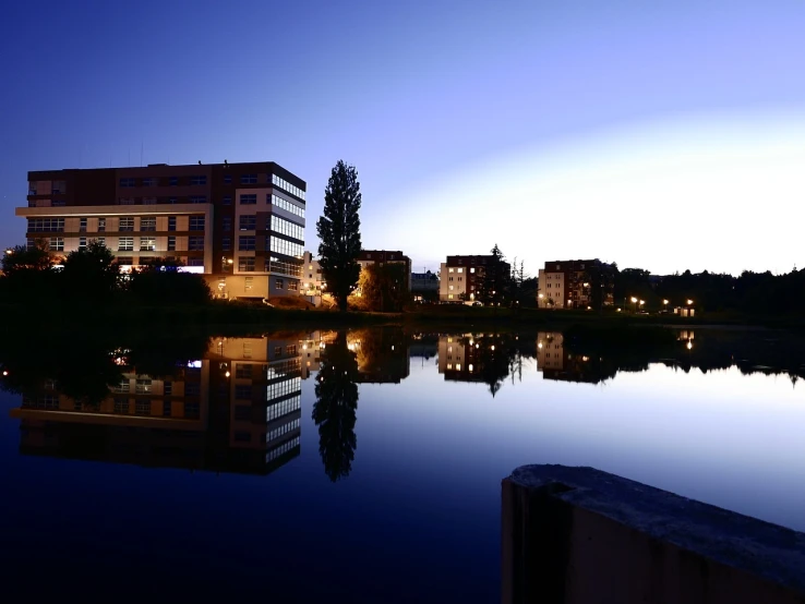 a body of water with buildings in the background, by Mathias Kollros, flickr, summer night, hospital, lake reflection, gradins view