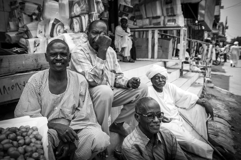 a group of men sitting next to each other, a black and white photo, by Ingrida Kadaka, the vibrant echoes of the market, nubian, smiling for the camera, stern expression