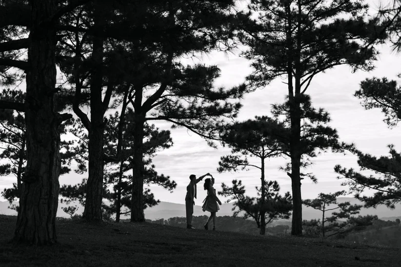 a couple of people that are standing in the grass, a black and white photo, by Tanaka Isson, hill with trees, couple dancing, pine trees, shot on sony alpha dslr-a300