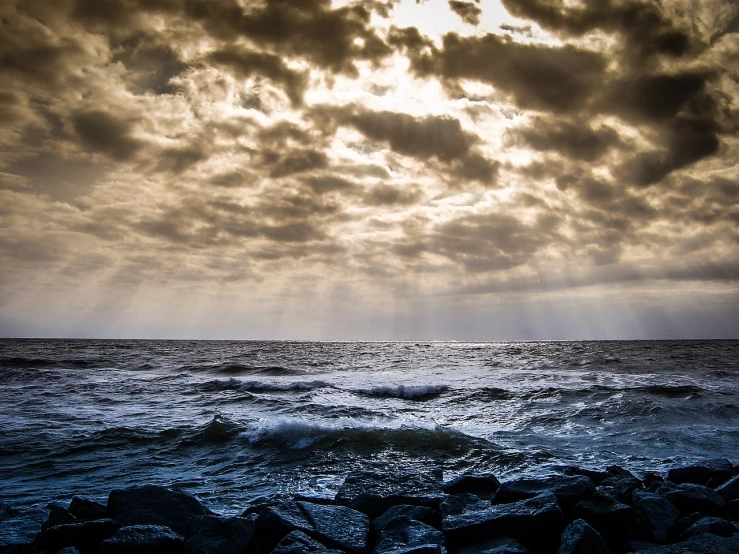 the sun shines through the clouds over the ocean, a picture, by Jan Konůpek, rough seas in background, photostock, rocky shore, journalism photo